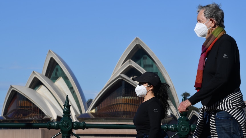 A man and a woman wearing masks walk past the Sydney Opera House. 