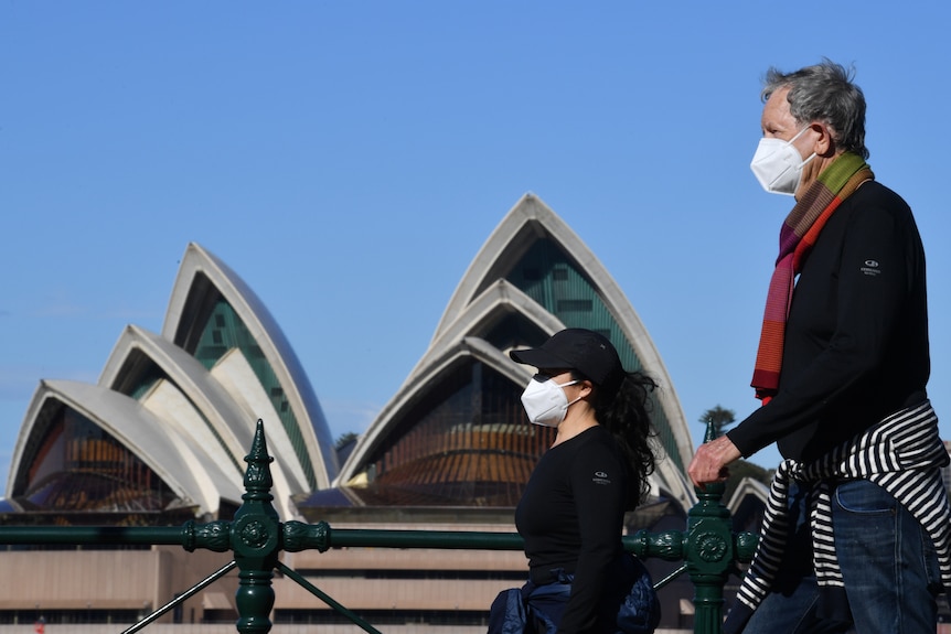 A man and a woman wearing masks walk past the Sydney Opera House. 