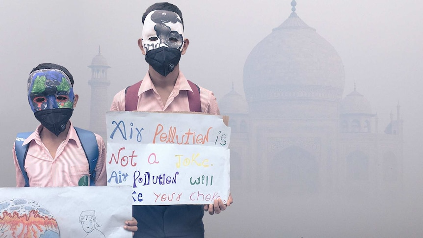 Indian boys wearing masks and holding signs with anti-pollution messages hand painted on them. Smoke obscures Taj Mahal behind.