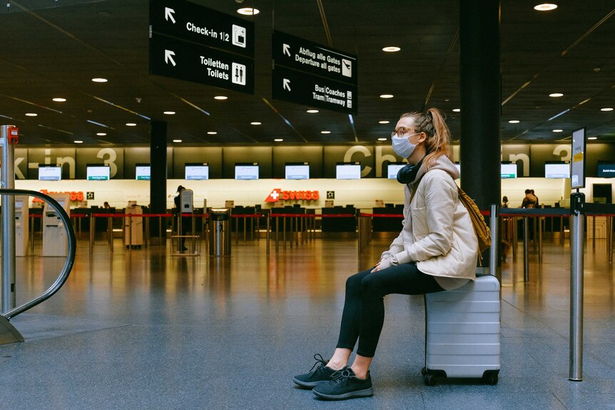 woman sits on suitcase in airport wearing mask
