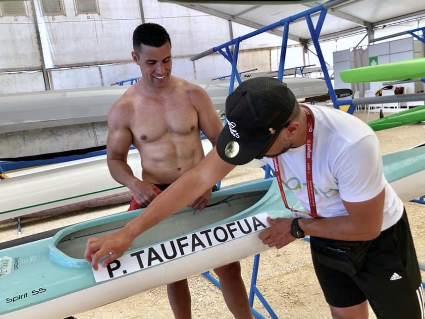 In a fabric-lined tent, Pita Taufatofua looks on, shirtless, as his coach places a sticker with his name onto a teal kayak.