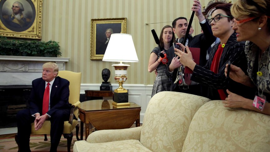 Donald Trump sits in a chair facing away from a group of journalists leaning.