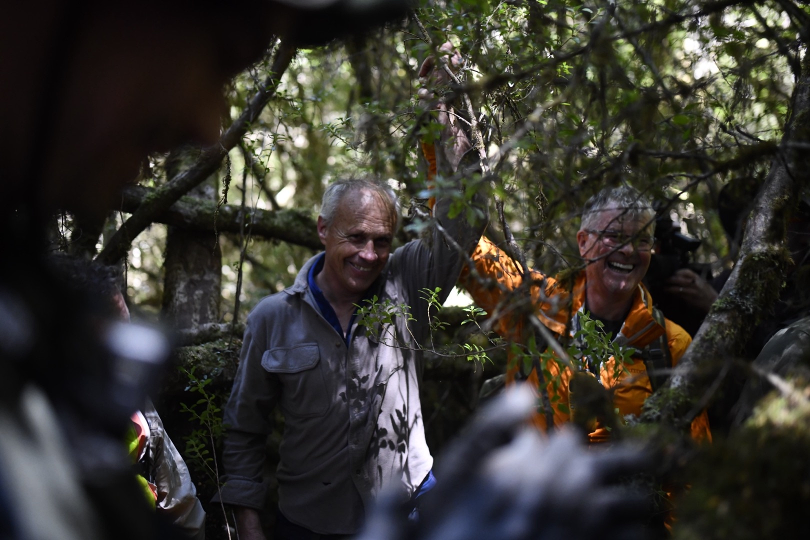 Dois homens de meia-idade e cabelos grisalhos sorriem em um matagal.