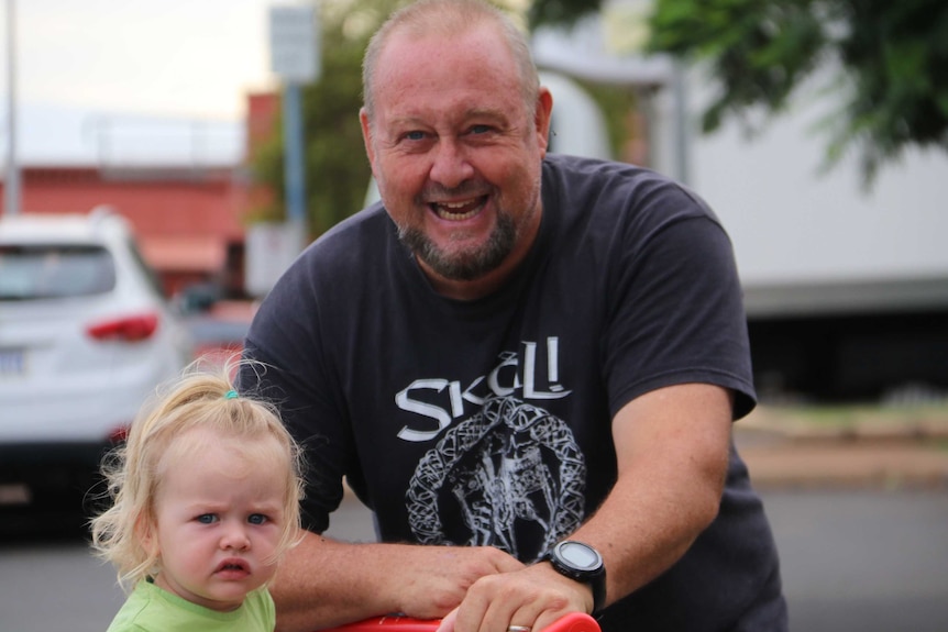 A man in a black t-shirt pushes a shopping trolley carrying a small girl outside a supermarket.