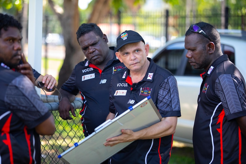 A coach of an Australian Rules football team coaches his players, holding a whiteboard