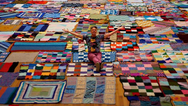 Ryk Goddard sits on colourful knitted blankets with arms stretched