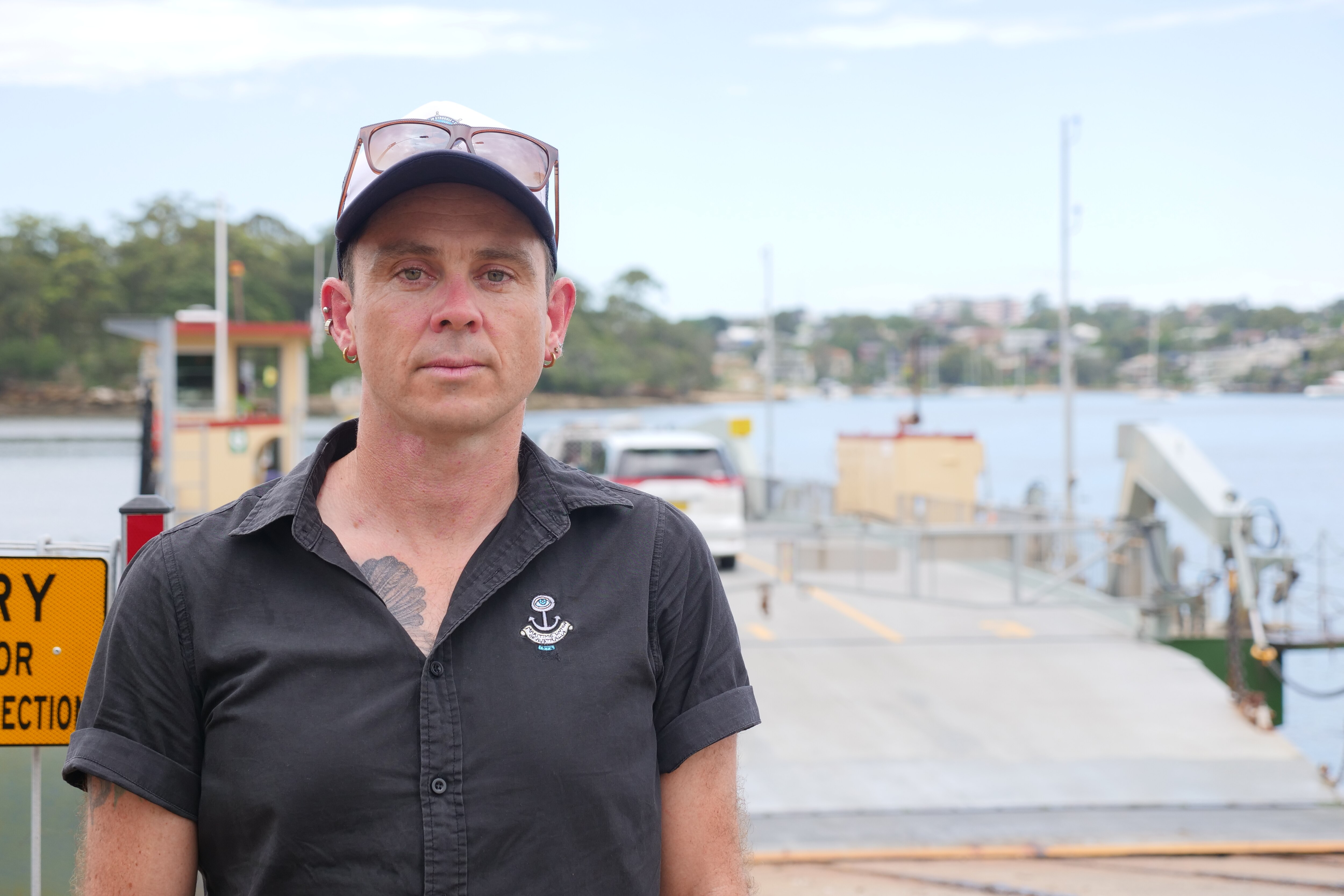 A man looks neutrally standing in front of water and a ferry