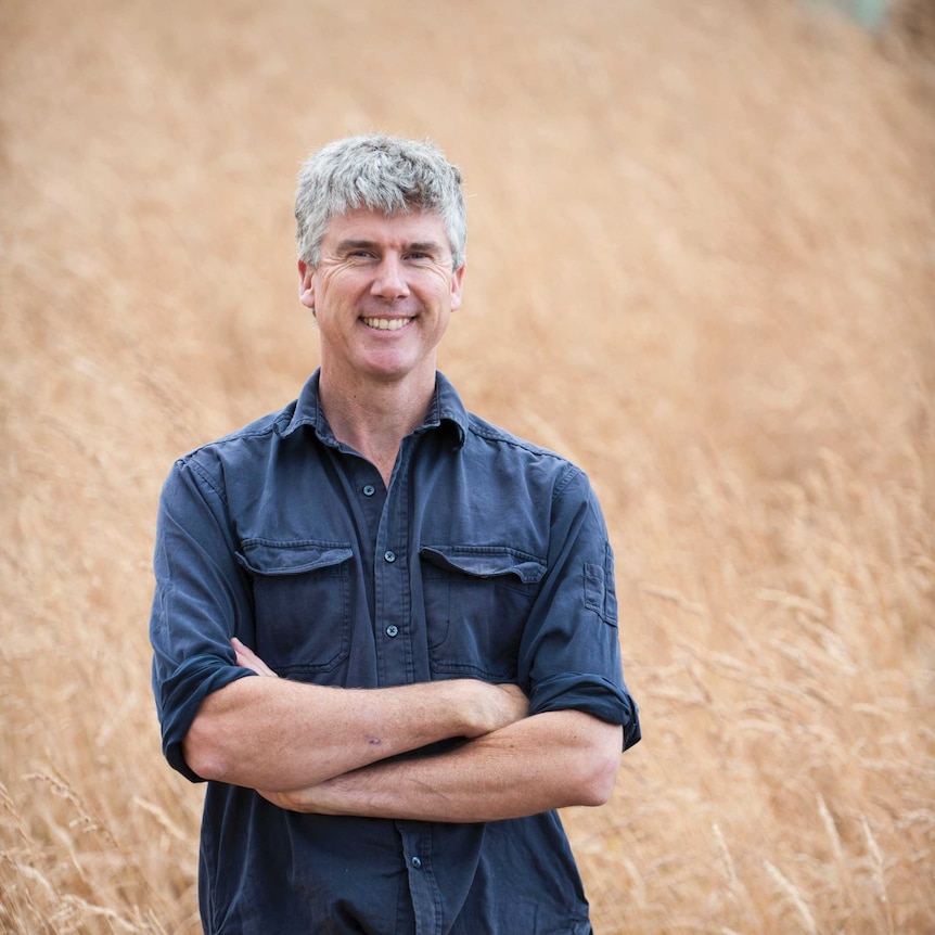 Matthew Evans is standing in a field of wheat and smiling, his arms are crossed in front of his chest.