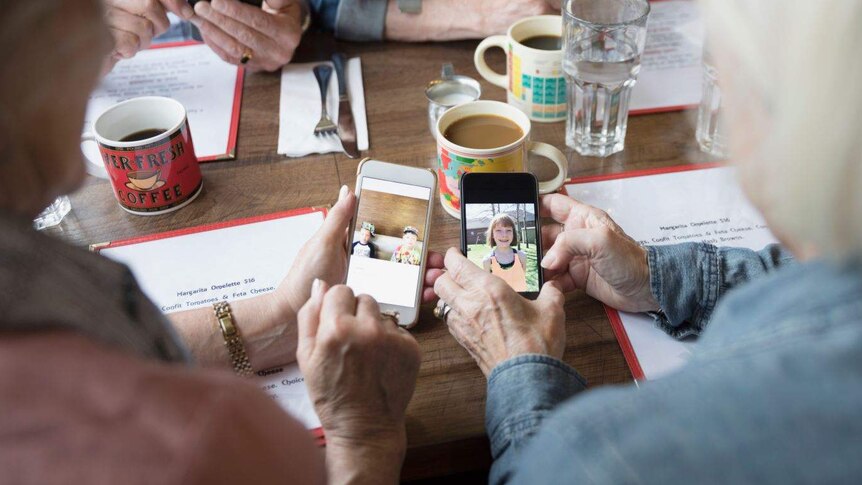 At a table covered in glasses of water and mugs of coffee, older hands hold smartphones on which are photos of children.
