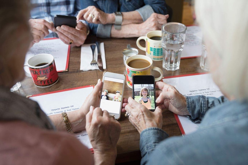 At a table covered in glasses of water and mugs of coffee, older hands hold smartphones on which are photos of children.