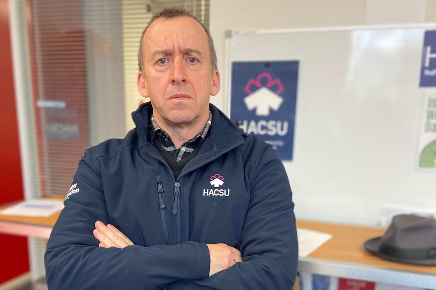 A man wearing a blue jacket stands with his arms crossed in an office with a serious expression.