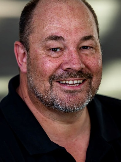 A bald man with short, light beard, wearing a dark polo shirt, smiling broadly.