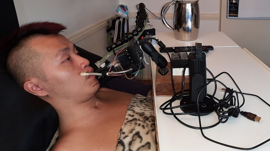 A young man lies in bed, he is using a mouth operated device to play video games.