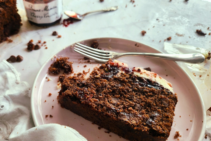 A slice of Ella Rossanis' chocolate cherry loaf on a light-pink plate, with the loaf and jam in the background.