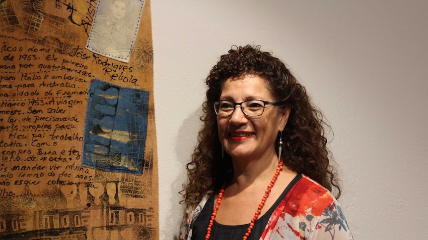 Artist Philomena Hali stands in front of an artwork, a long scroll covered in imagery and writing in Portugese.