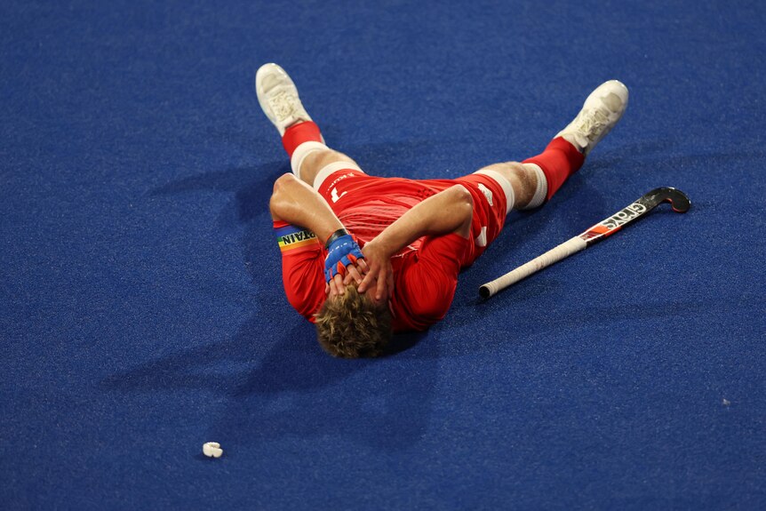 An England hockey player in red lies on the astroturf with his hands over his face and his hockey stick lying next to him.