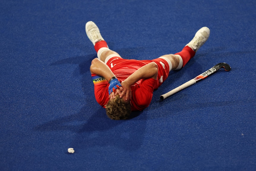 An England hockey player in a red suit lies on the astroturf, covering his face with his hands and holding a hockey stick by his side.