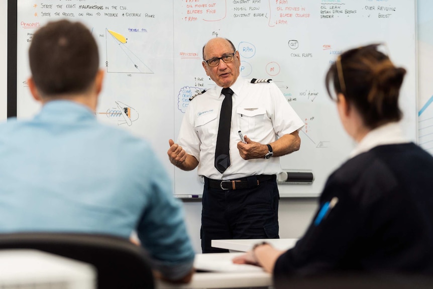 A flying instructor stands at the front of a classroom in front of a whiteboard talking to a man and a woman sitting down.