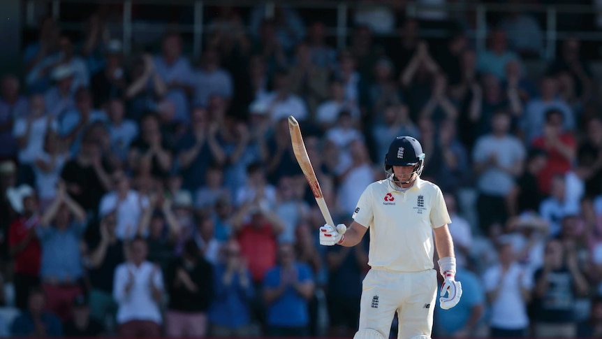 Joe Root holds his head down but holds his bat up in his right hand. The crowd applauds in the background.