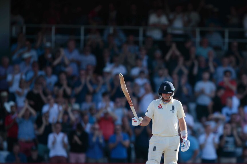Joe Root holds his head down but holds his bat up in his right hand. The crowd applauds in the background.