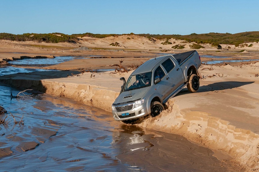 A four wheel drive ute driving down a small steep sandy bank into a stream on a beach