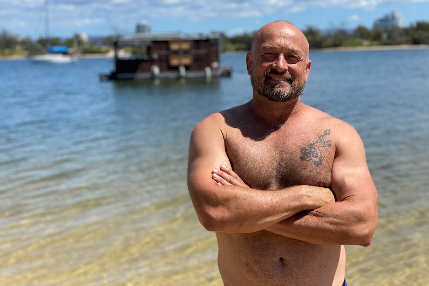 Shirtless man standing with his arms crossed in front of a floating brown houseboat. 