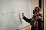 A young woman stands at a whiteboard, wearing a headscarf.