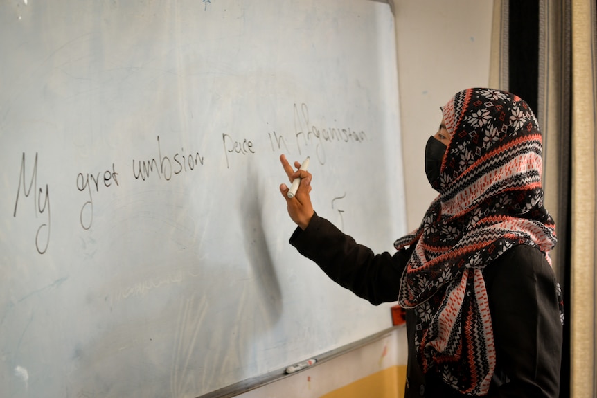 A young woman stands at a whiteboard, wearing a headscarf.