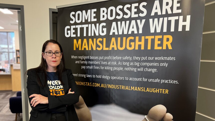 Jess Munday stands next a campaign sign that says 'some bosses are getting away with manslaughter'