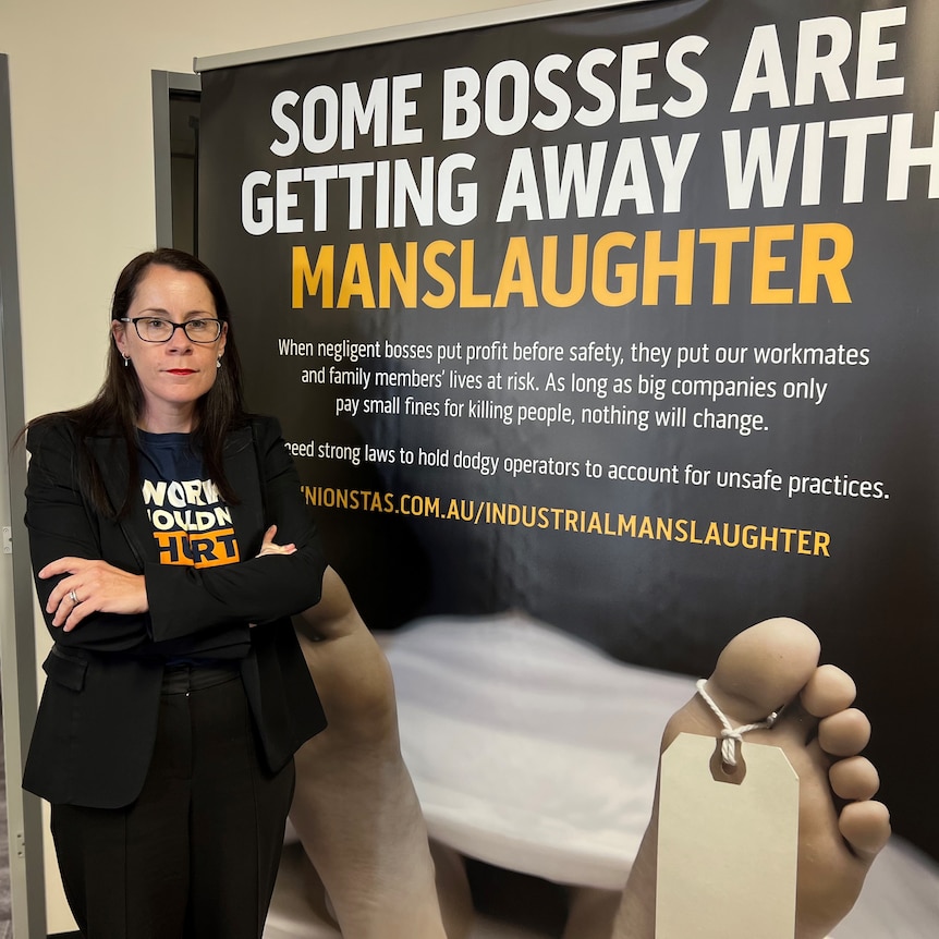 Jess Munday stands next a campaign sign that says 'some bosses are getting away with manslaughter'