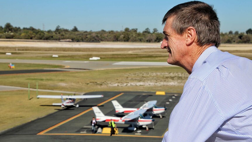 A man in a blue shirt looks out over an airport runway where several light aircraft are parked.