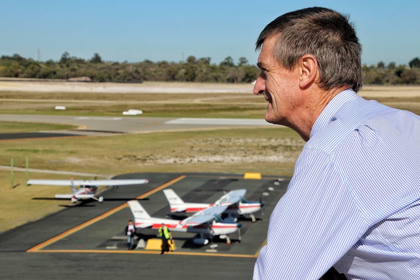 A man in a blue shirt looks out over an airport runway where several light aircraft are parked.