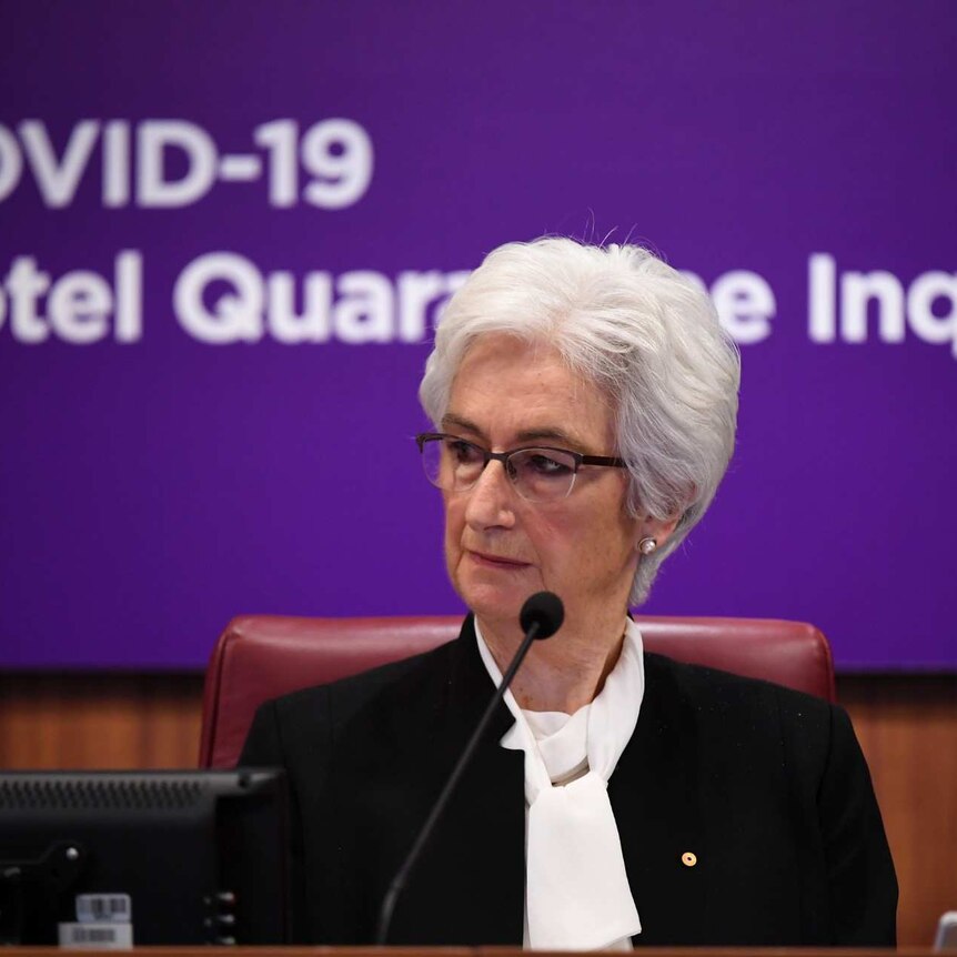 Jennifer Coate sits behind a bend with the words "COVID-19 Hotel Quarantine Inquiry" on the wall behind her.