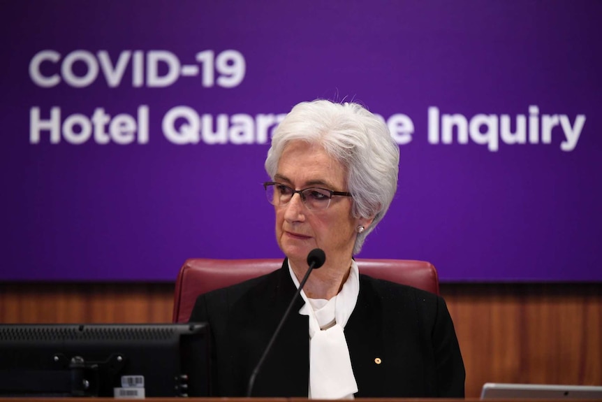 Jennifer Coate sits behind a bend with the words "COVID-19 Hotel Quarantine Inquiry" on the wall behind her.