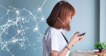 A young woman looks at a mobile phone while standing at a desk.