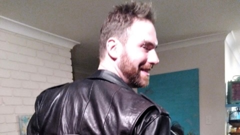 A man with his back towards the camera grabs his leather jacket which reads "unlovable". You can see his profile.