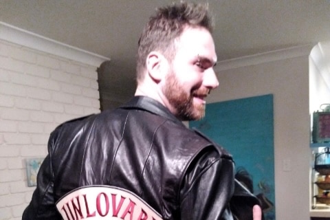 A man with his back towards the camera grabs his leather jacket which reads "unlovable". You can see his profile.