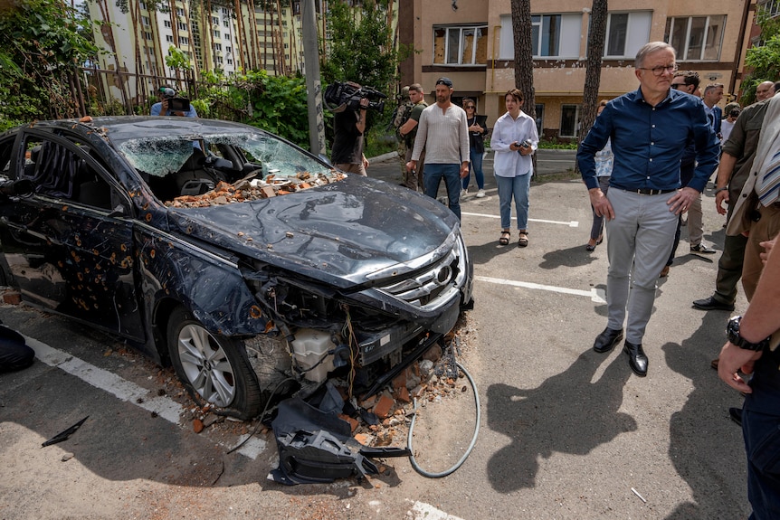 Albanese stands with his hands on his hips beside a bombed car, surrounded by locals and press.