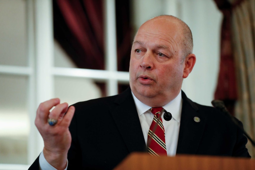 A bald man in a black suit gestures with his hands.