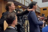 A young white woman reaches out and touches the shoulder of a young Chinese man amid a crowd of reporters.