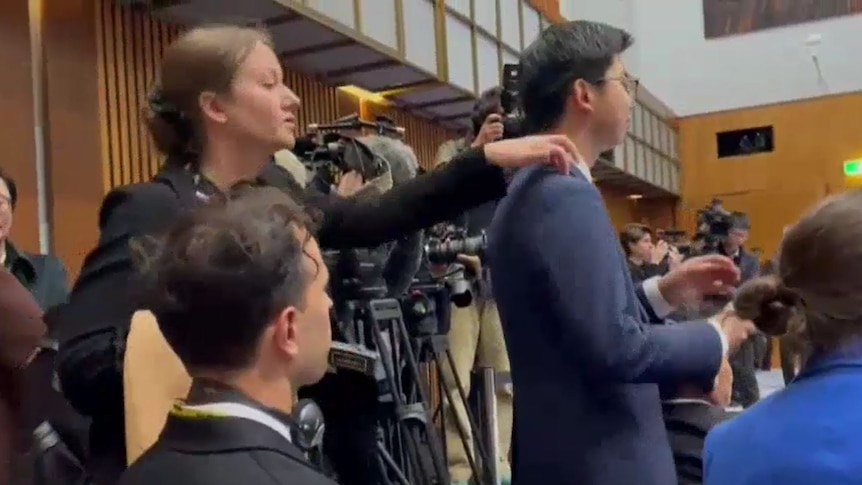 A young white woman reaches out and touches the shoulder of a young Chinese man amid a crowd of reporters.