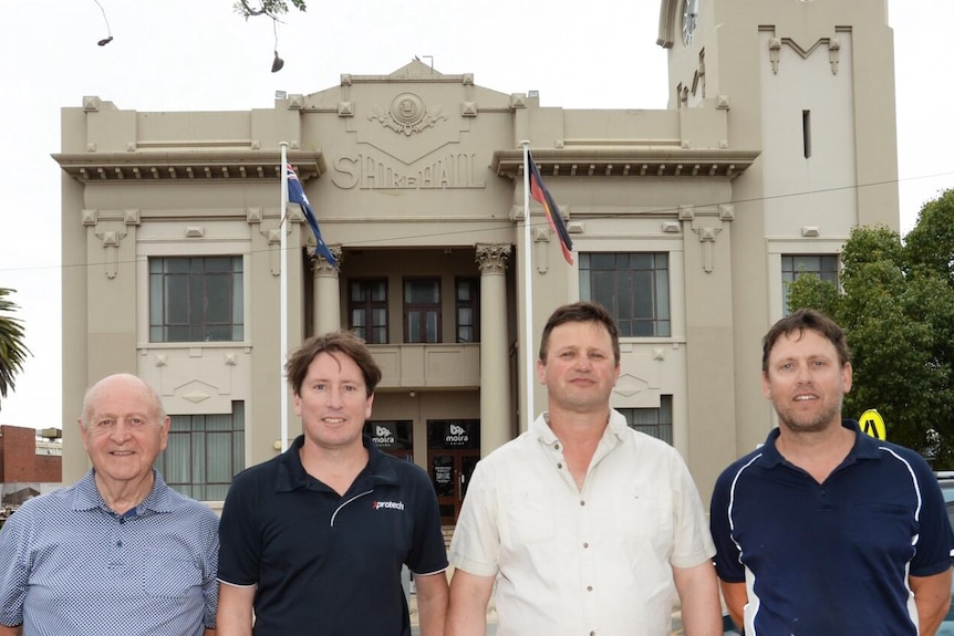 A group of men in collared shirts stand in front of a municipal building. 