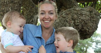 young woman stands holding two young children who are interacting with each other and mum is smiling looking at the camera