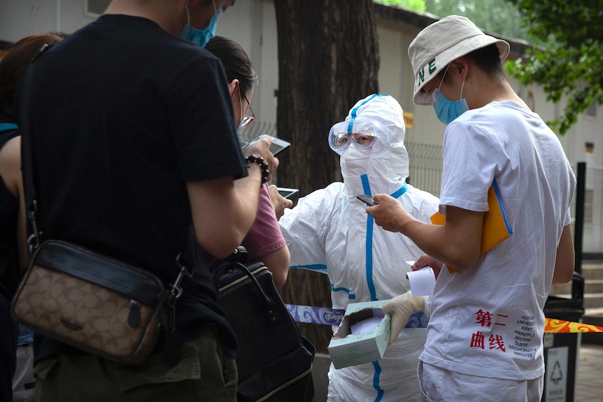 A health worker in full PPE gear talks to a group of people holding their phones