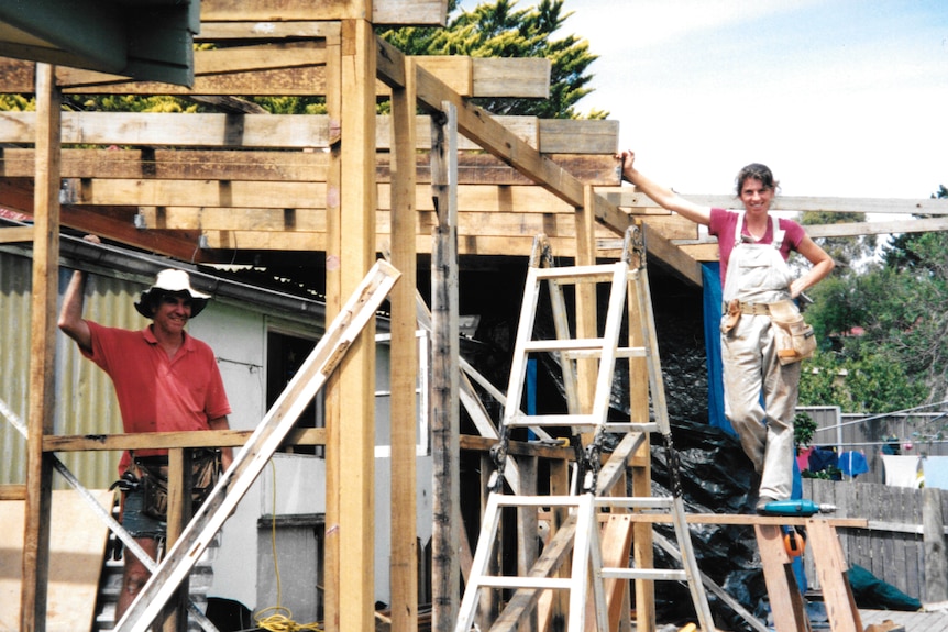 Two people stand on ladders and smile during the construction of a house.