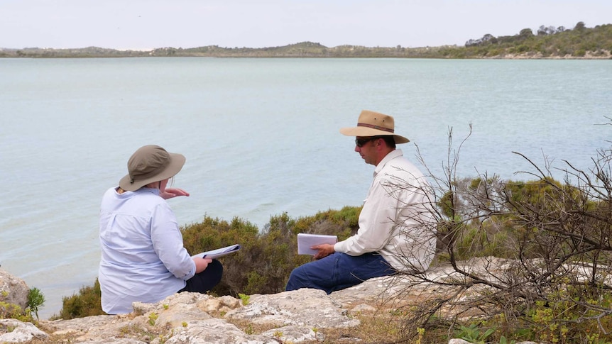Woman on left and man on right perched on ledge with sea outlook and notebooks