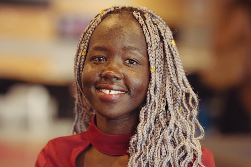 A young African-Australian woman with long, blonde braids and a smart red top smiles brightly at the camera.