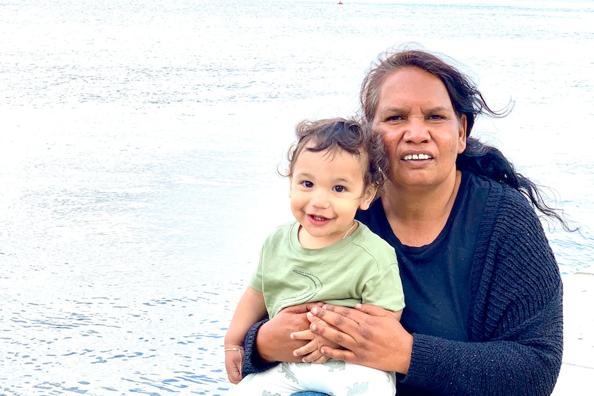 Megan is sitting by the water with a baby in her arms smiling at the camera