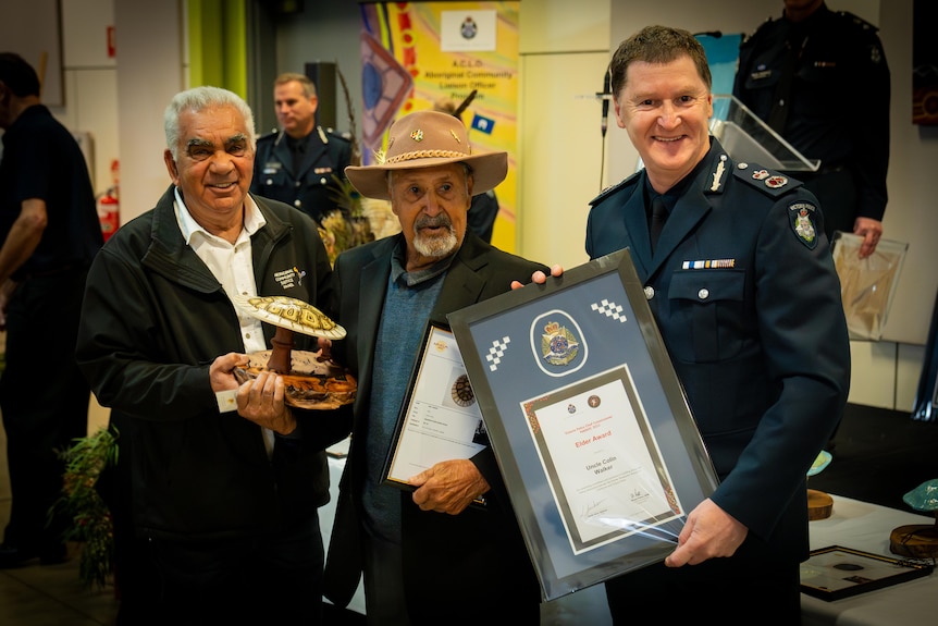 Three men standing side by side with the middle one holding an award trophy in one hand and a framed award in another