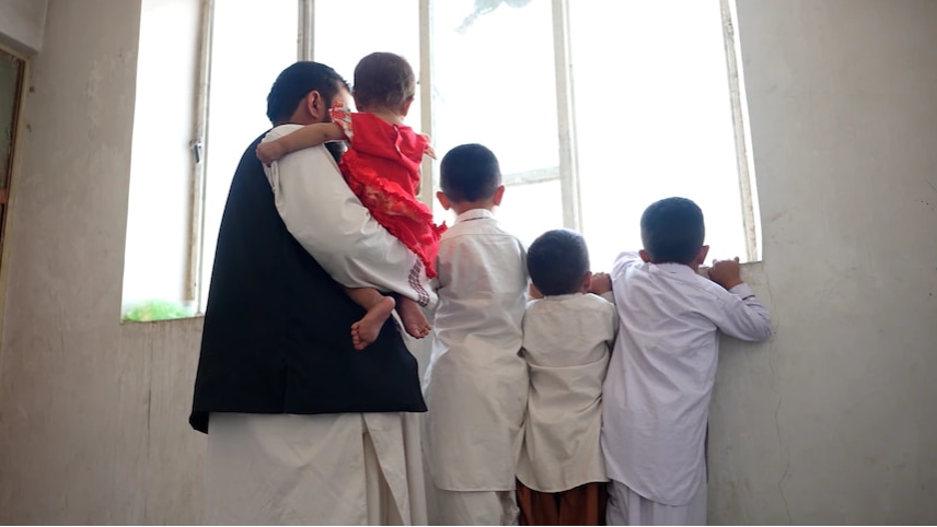 The back of a man holding a baby stands with three young children looking out a window